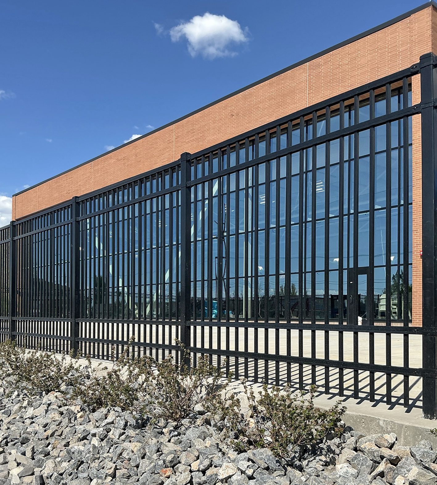 Modern brick and glass building behind a black metal fence, with rocks and bushes in the foreground under a blue sky.