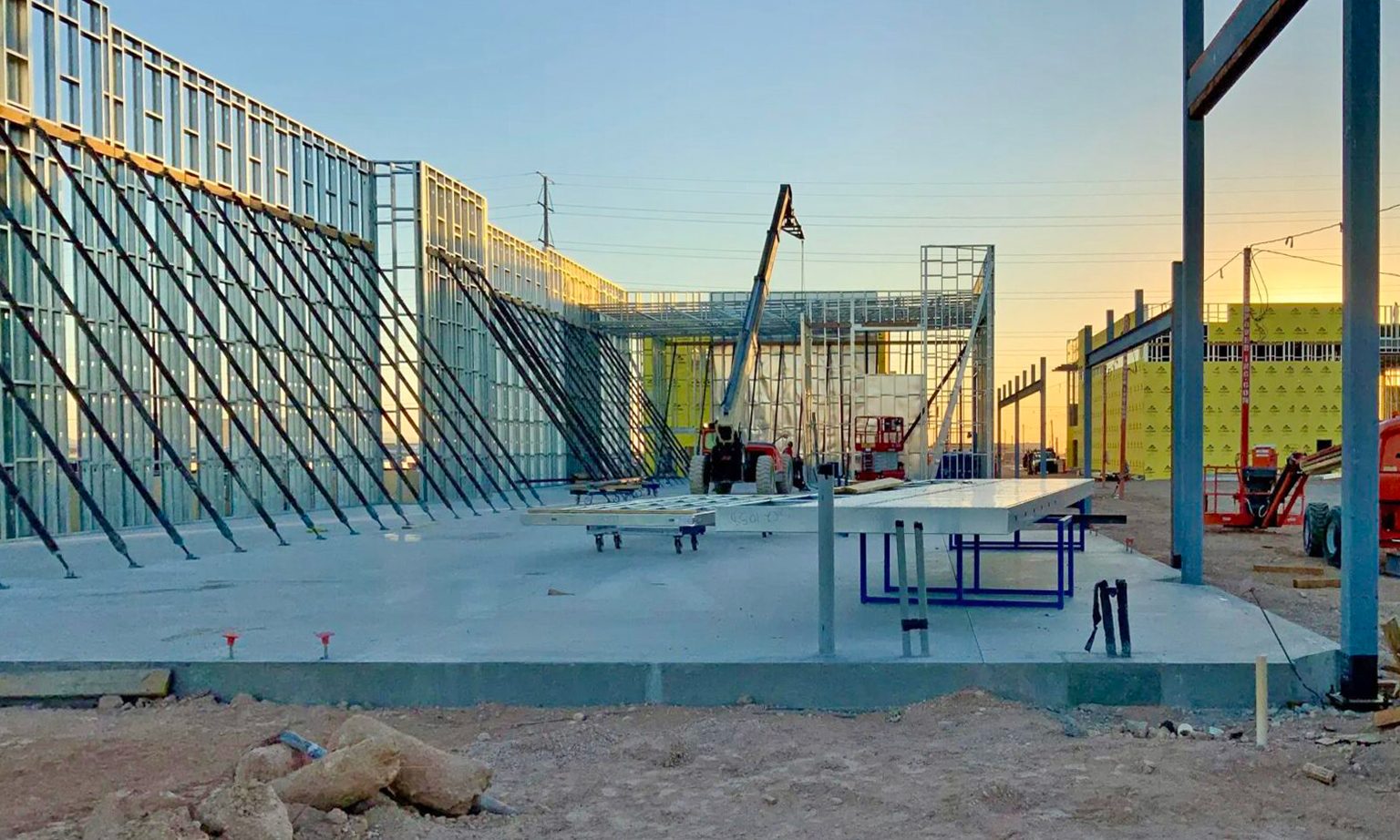 A building under construction with metal wall framing, concrete foundation, and machinery at sunset.