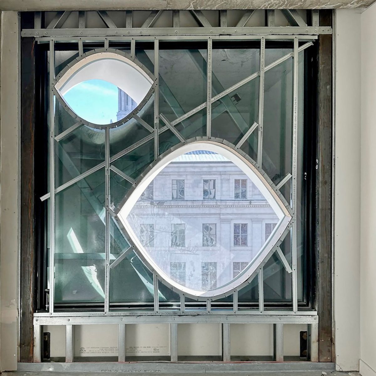 Abstract architectural window with metal framing, creating two eye-shaped openings revealing sky and a building.