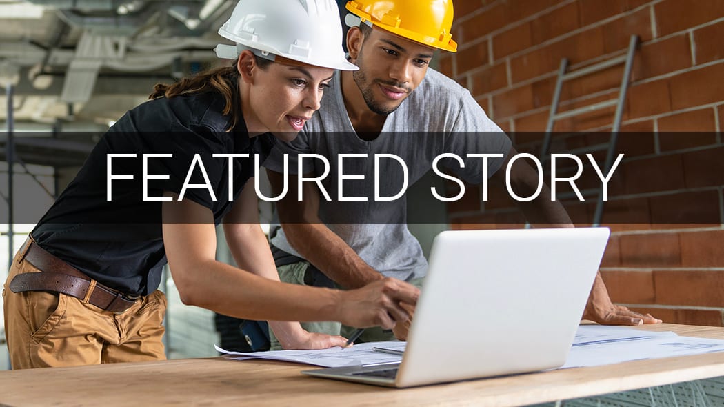 Two construction workers in hard hats review blueprints on a laptop at a building site.