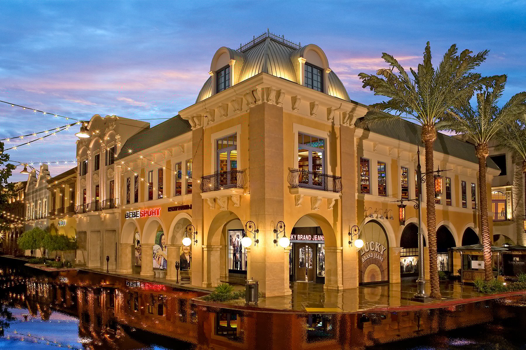 An ornate, illuminated shopping street building with palm trees and reflections on a wet evening.
