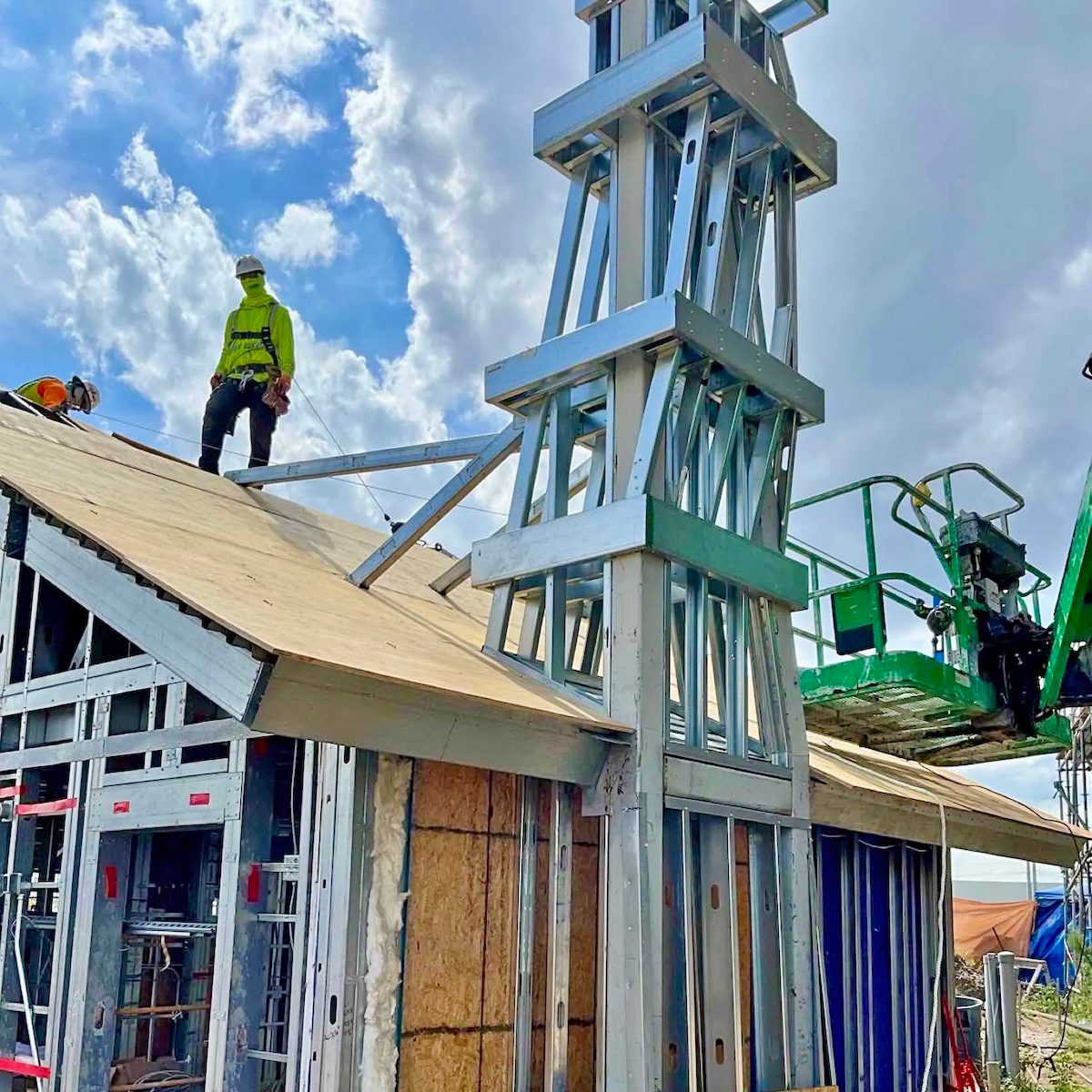 Steel frame house under construction with workers on the roof and a complex metal tower rising.