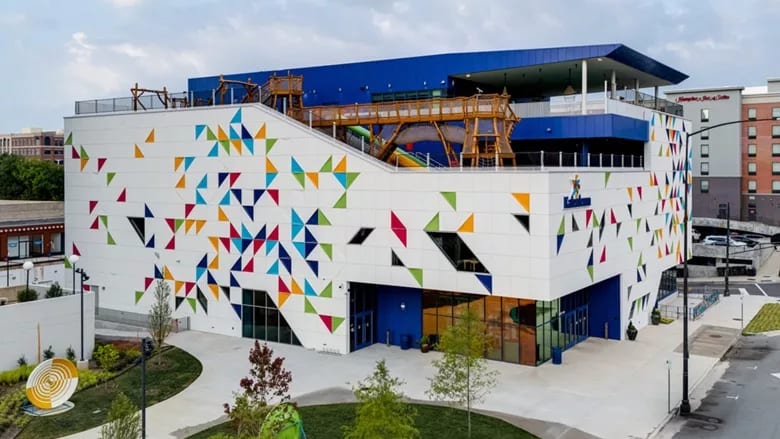 White building adorned with colorful triangles, a blue roof, and an elevated wooden playground.