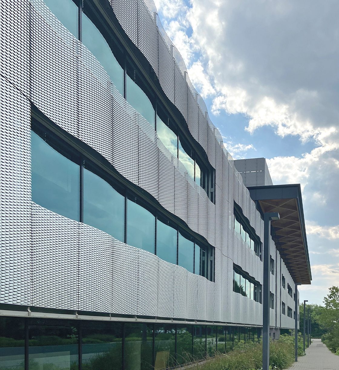 Modern building with a wavy, perforated silver facade, large reflective windows, and a clear sky with clouds.