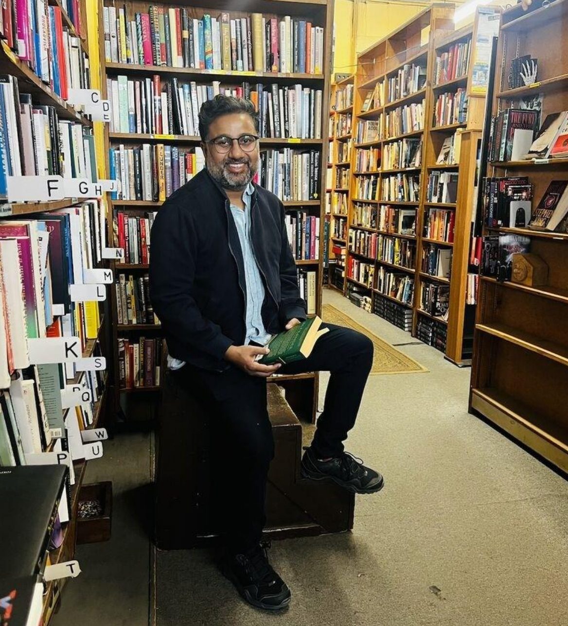 Dr. Pradyumna (Prady) Gupta. Smiling man sits on a stool in a bustling bookstore, holding a green book.