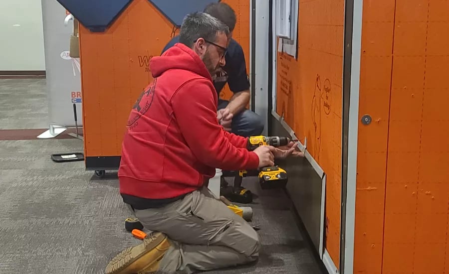 Man in red hoodie kneels, drilling an orange panel. Another man is behind him.