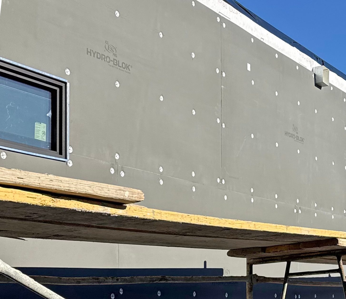 Building exterior under construction with gray HYDRO-BLOK insulation panels, a window, and scaffolding.