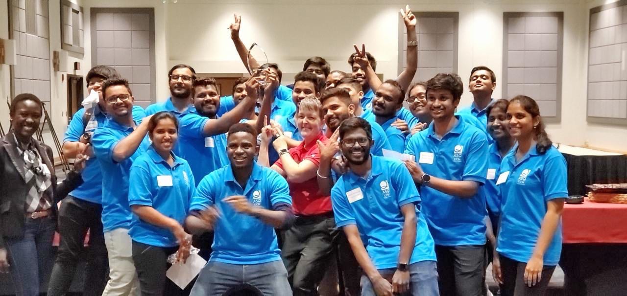 A large, diverse group of people in blue shirts and one woman in red, celebrating with a trophy.