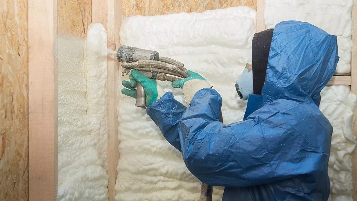 Person in protective suit spraying insulation foam on a wooden wall.