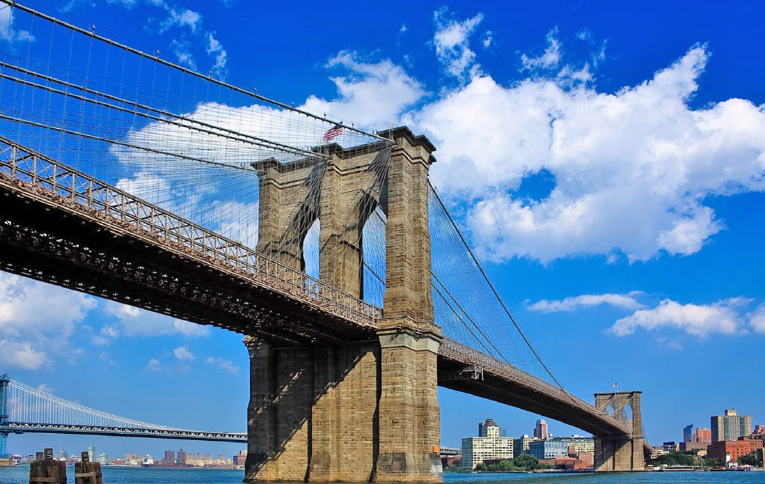 Brooklyn Bridge's majestic stone towers and cables spanning the East River, under a bright blue sky with clouds.