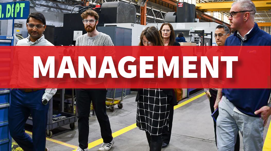 Group of people, some wearing safety glasses, walking through a factory with machinery in the background.