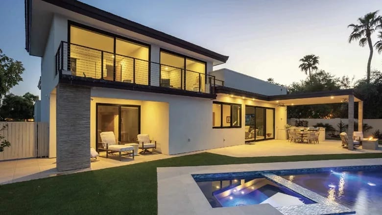 Modern luxury house exterior with illuminated swimming pool, covered patio dining area, and balcony at dusk.