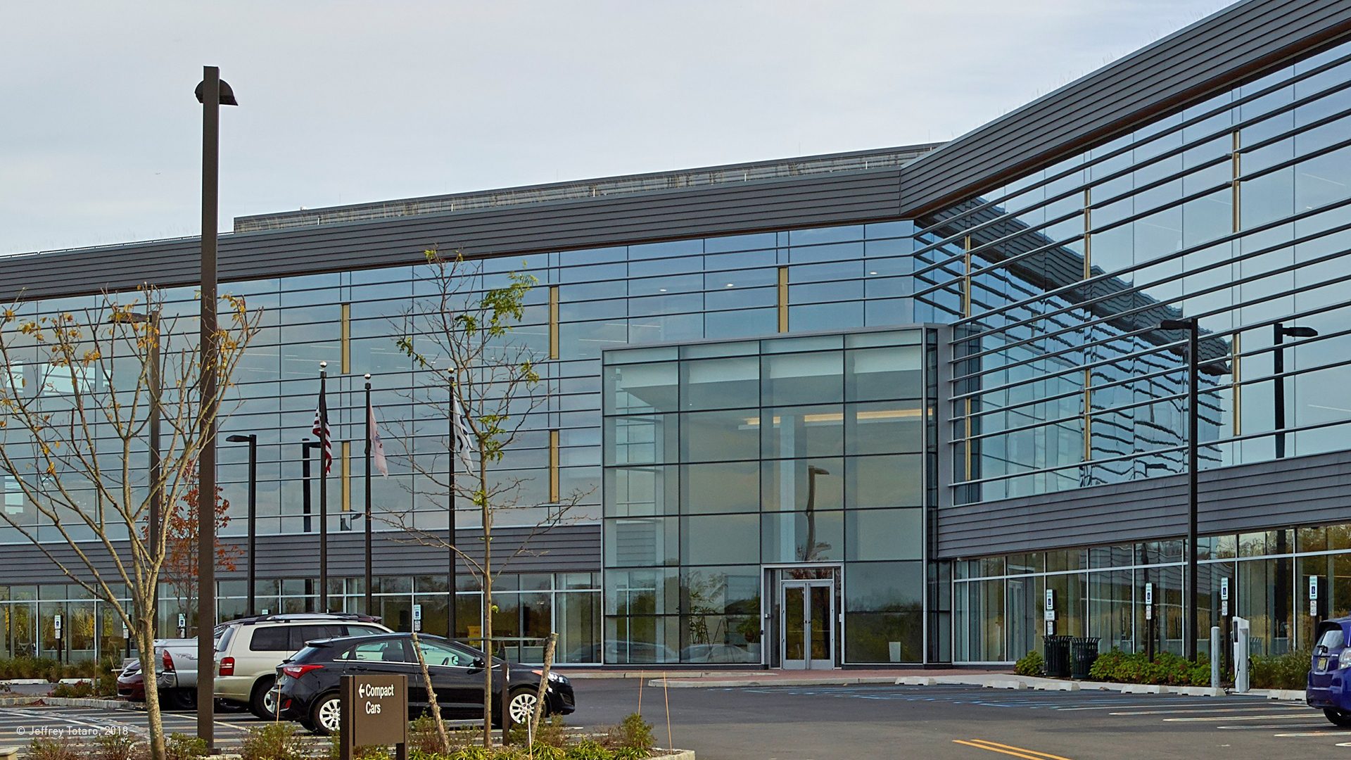 Modern glass building, parking lot with cars, trees, and flags.