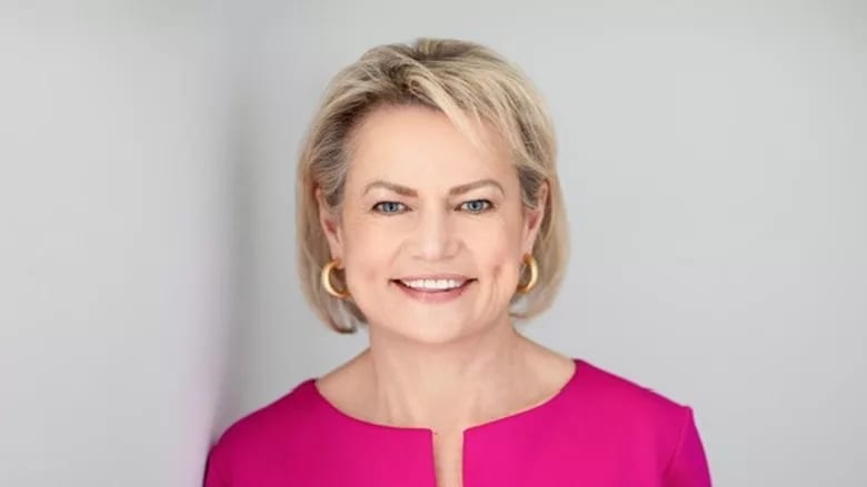 A smiling woman with blonde hair, blue eyes, and gold hoop earrings, wearing a bright pink top.