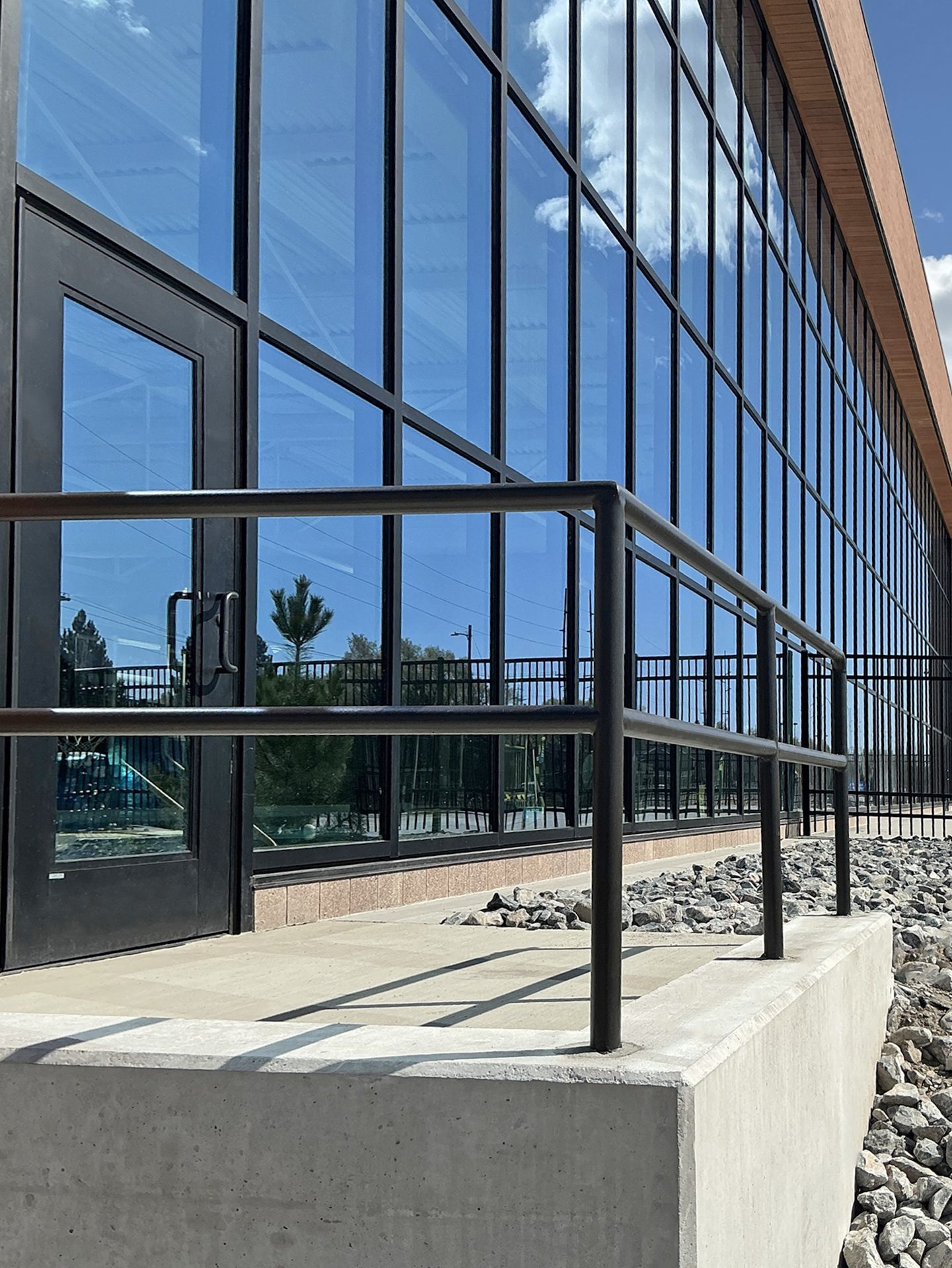 Modern building with reflective glass facade, sky/cloud reflections, black railing, and concrete platform.