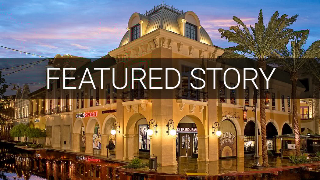 Illuminated shopping center at dusk with reflections, featuring ornate architecture and palm trees.