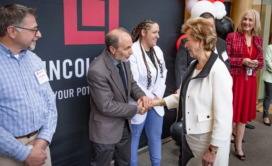 A group of people attending an event, with two individuals shaking hands in the foreground. A banner is visible in the background.