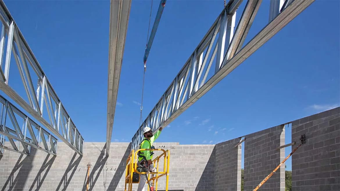 Construction worker in a lift guides a large metal truss into place on a building structure under a blue sky.