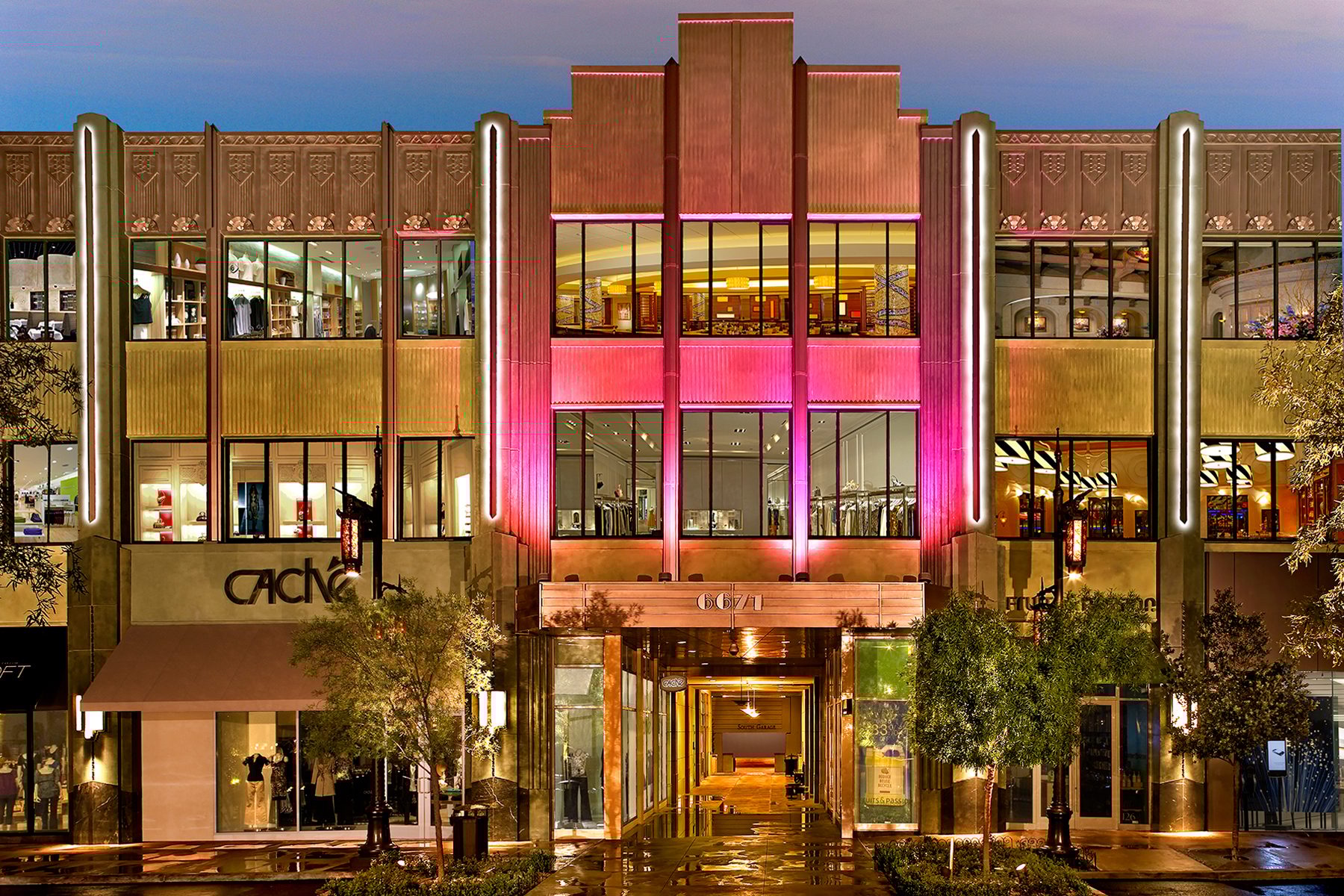 Illuminated Art Deco building facade at night, featuring retail stores, pink and gold lighting.