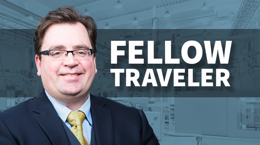 Smiling man in suit, glasses, and "FELLOW TRAVELER" text on blurred industrial background.