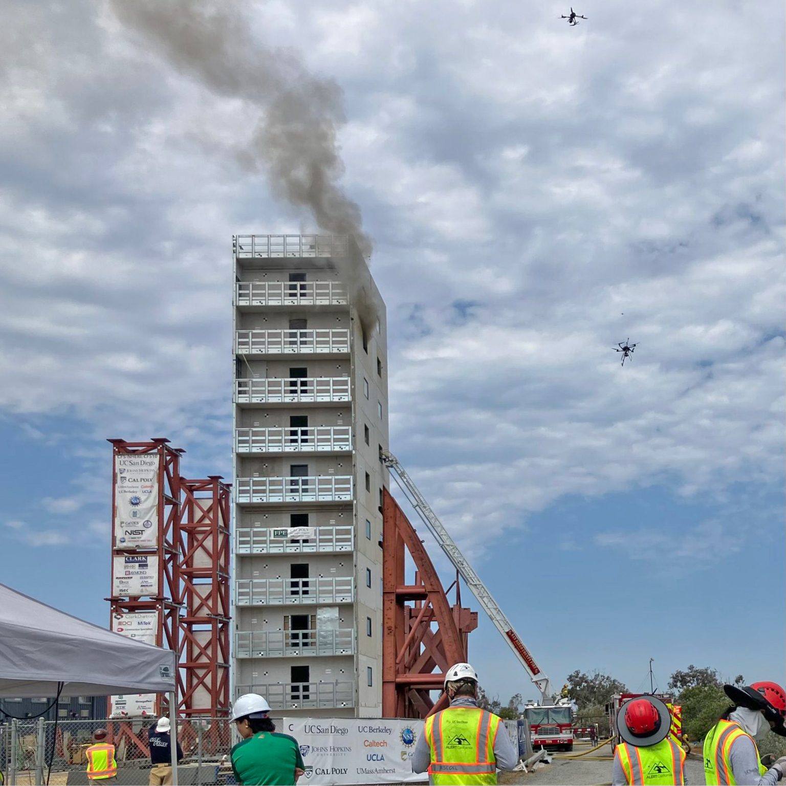 A burning multi-story building with smoke, two drones, and people observing a fire drill or test.