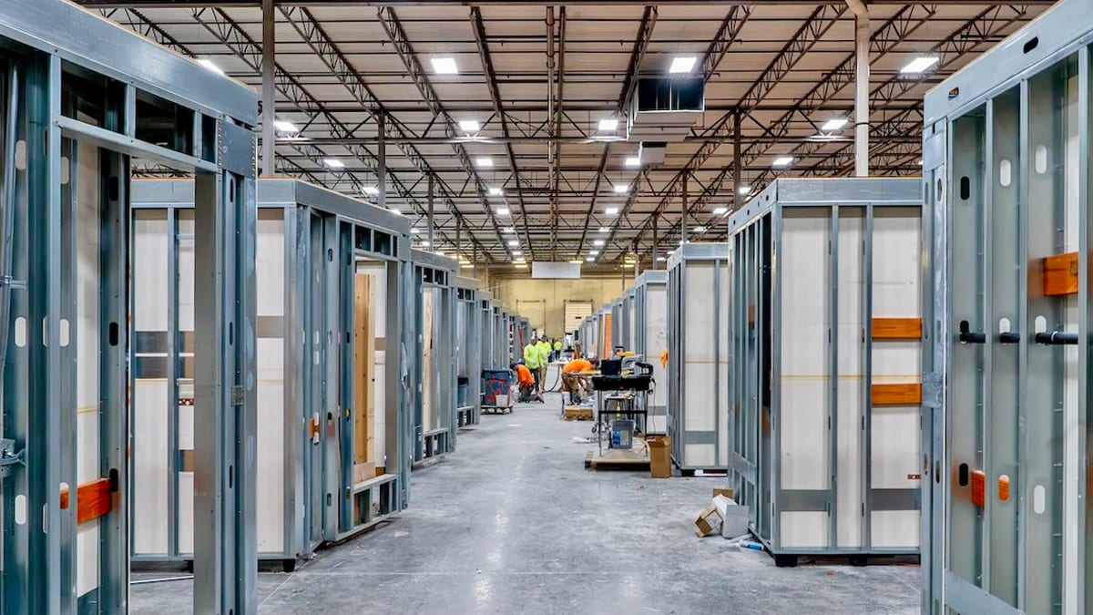 Workers assemble modular metal frame units in a large industrial warehouse during construction.