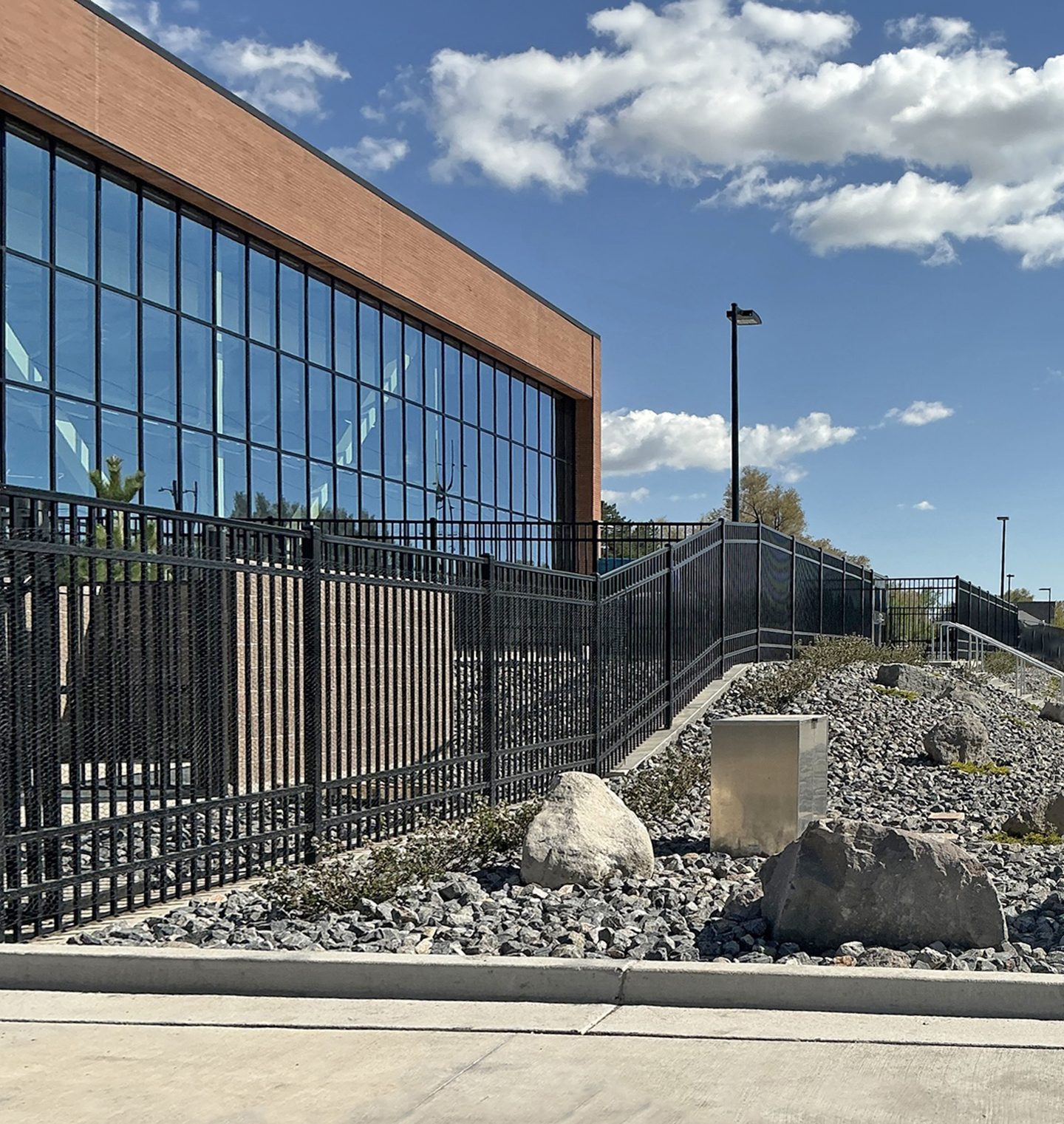 A modern building with a glass facade and brick top, flanked by a black fence and rocky landscaping.