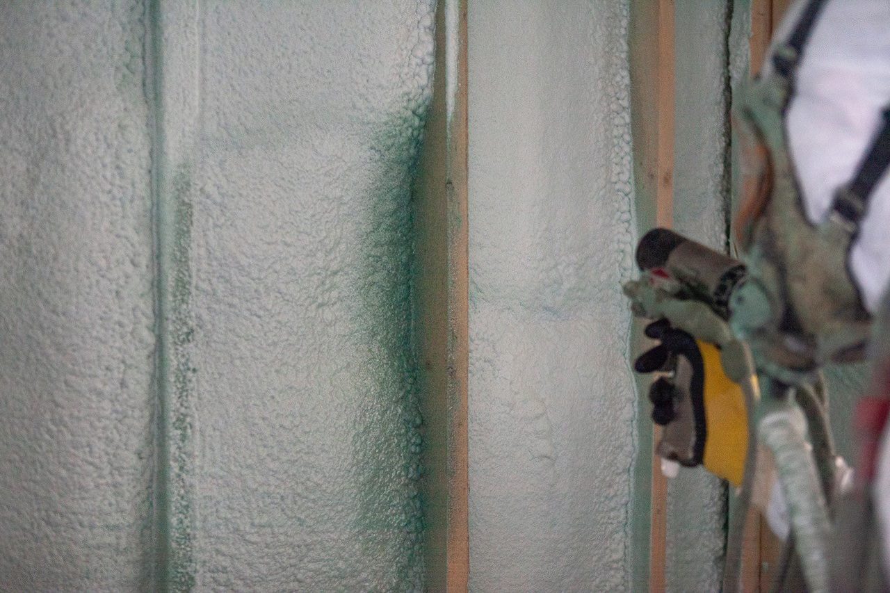 Hand spraying green foam insulation onto a wall between wooden studs.