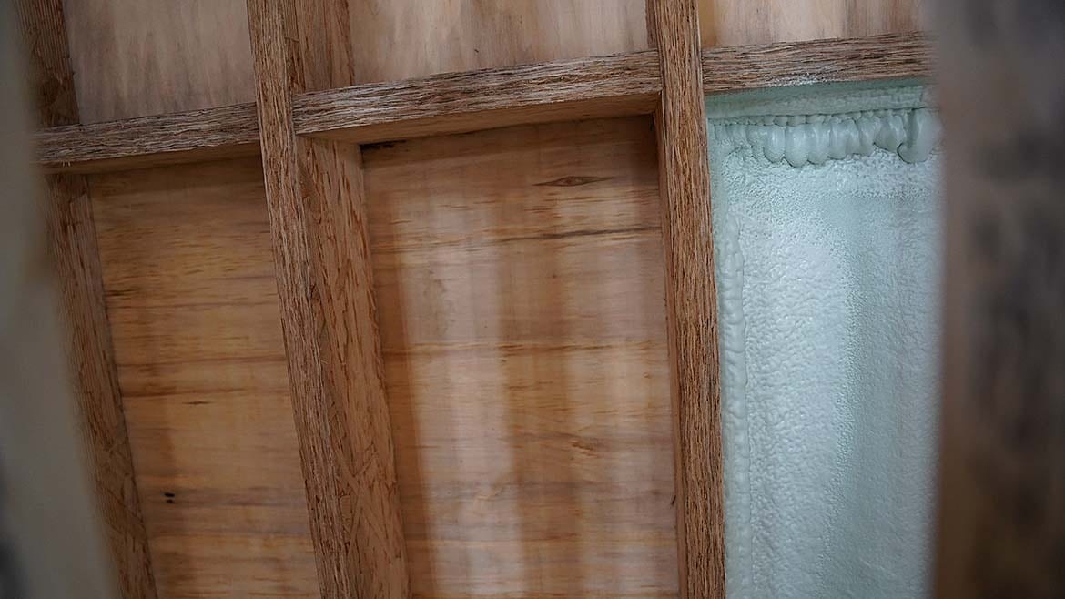 Wood framing, plywood, and light blue spray foam insulation filling a cavity.