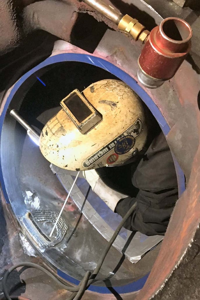 A welder in a large industrial component performs a weld, wearing a protective helmet and gloves.