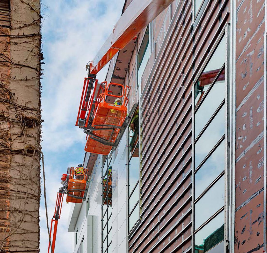 Two workers in orange boom lifts work on the facade of a modern building next to an old brick wall.