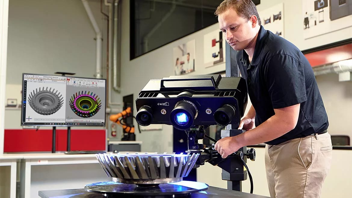 A man operates a 3D scanner to digitize a mechanical part, with CAD models displayed on a computer monitor.