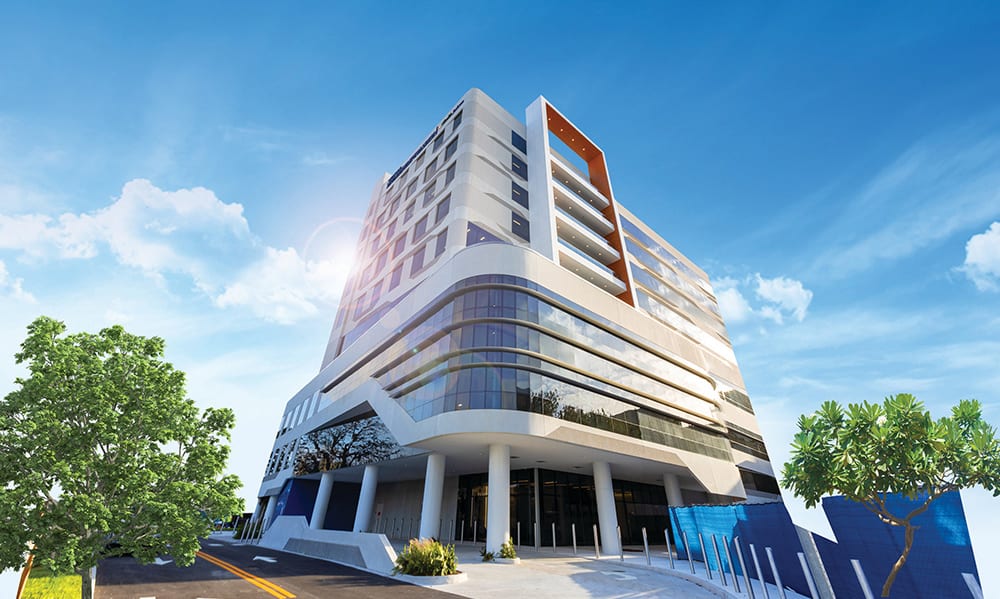 Modern high-rise building with glass facade, sun flare, and trees, under a bright blue sky.
