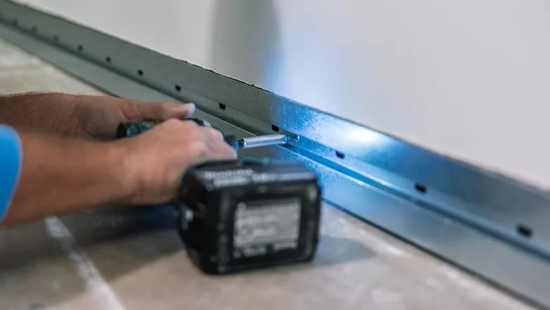 A person uses a cordless drill to fasten a screw into a metal track, illuminated by the drill's light.