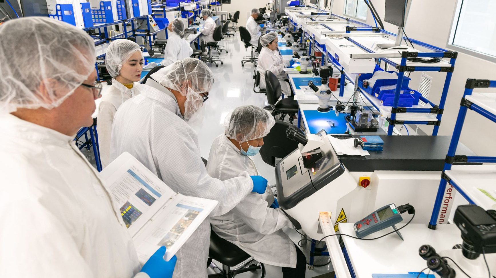 Technicians in protective gear work on a cleanroom production line with microscopes and other equipment.