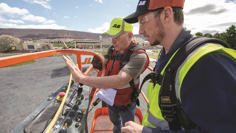 Two men in safety gear on a JLG aerial lift, one operates controls, with a factory in the background.