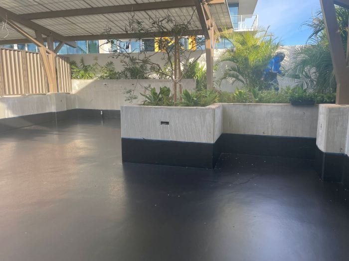 Newly waterproofed black patio floor with concrete planters, green foliage, and a person working in the garden.