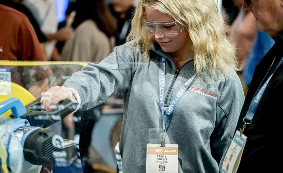 A young woman in safety glasses and a grey jacket operates a yellow and blue cutting machine at METALCON.