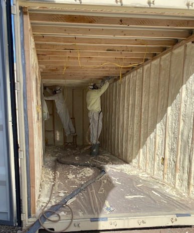 Two workers spray foam insulation inside a framed shipping container.
