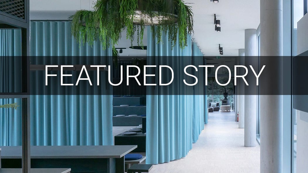 Modern office interior with blue acoustic curtains, hanging green plants, and stepped seating areas.