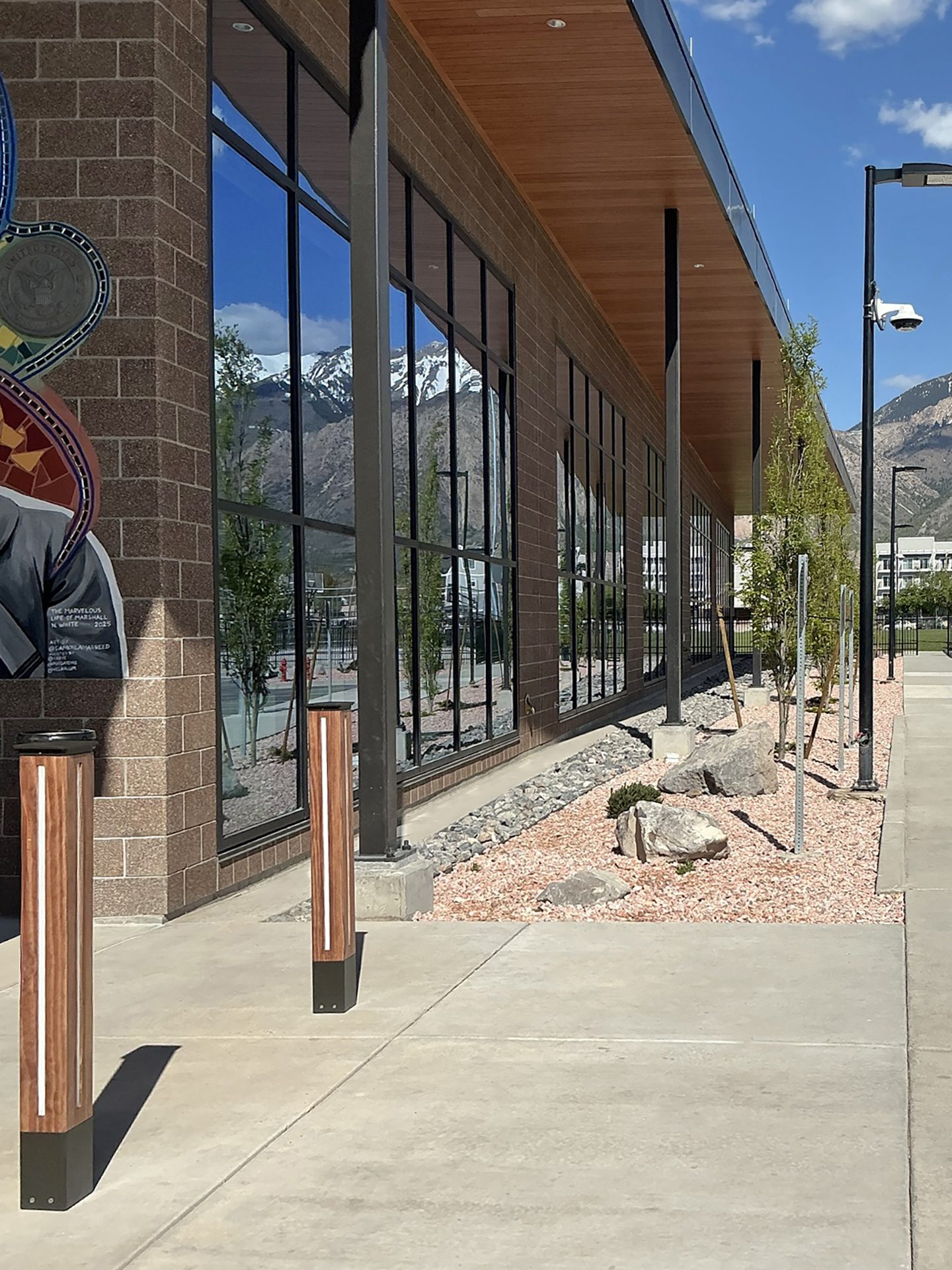 A modern brick building with large windows reflecting snowy mountains. Sidewalk with bollards and landscaping.