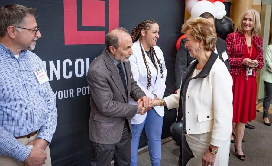 Two professionals shake hands at an event, with colleagues observing in the background.