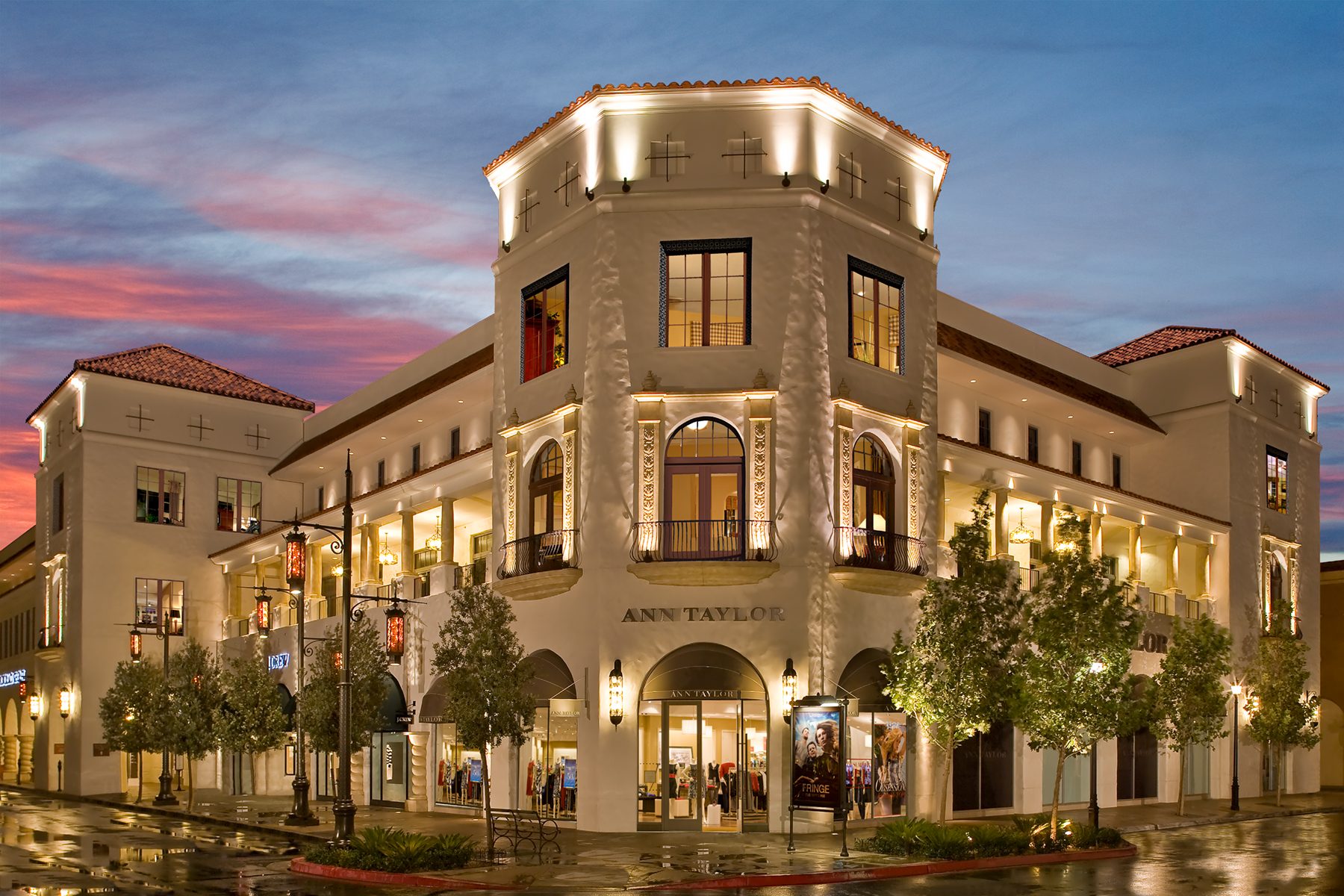 An illuminated Ann Taylor store building with Spanish-style architecture at dusk under a colorful sky.