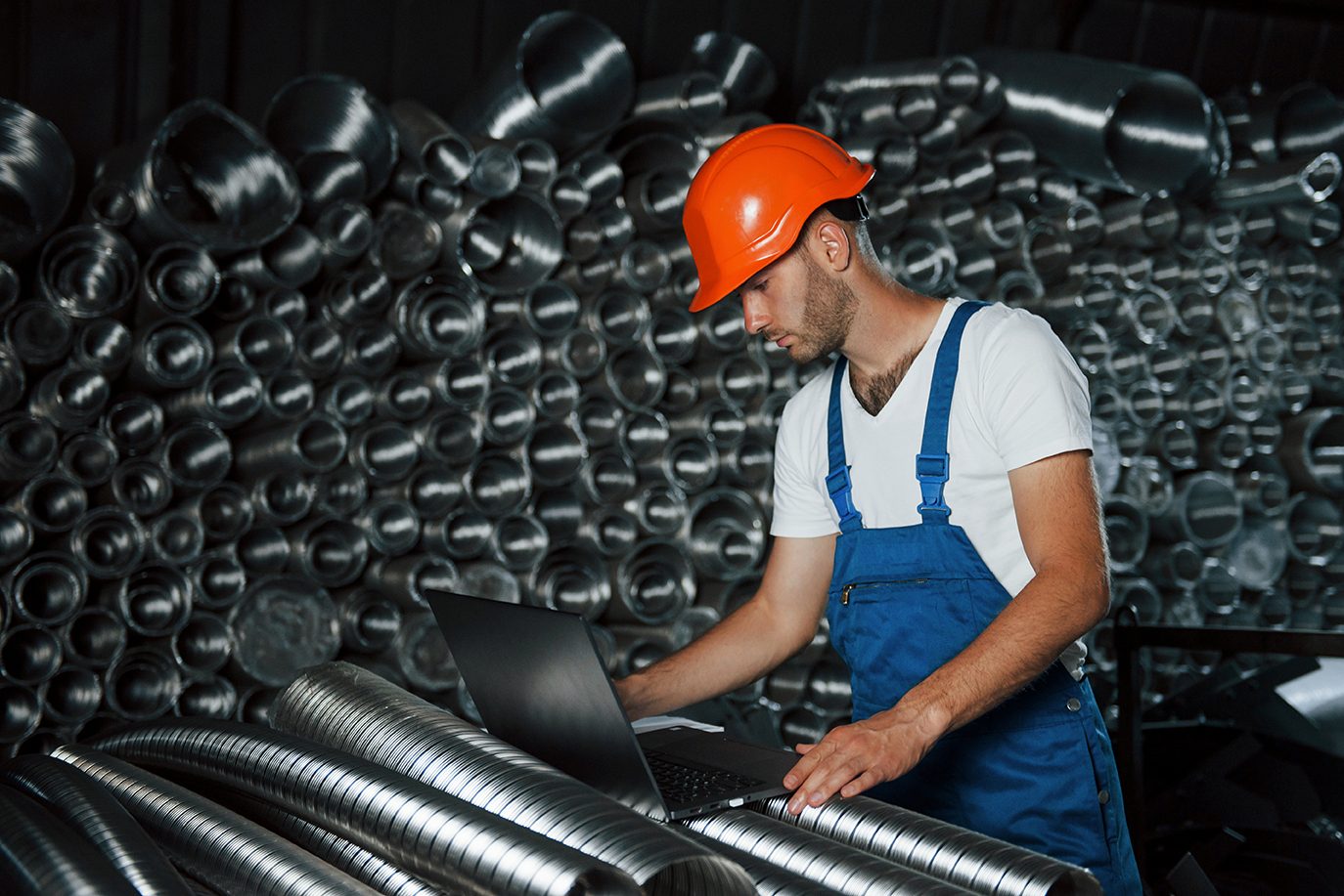 Man in hard hat and overalls uses laptop surrounded by metal ducts.