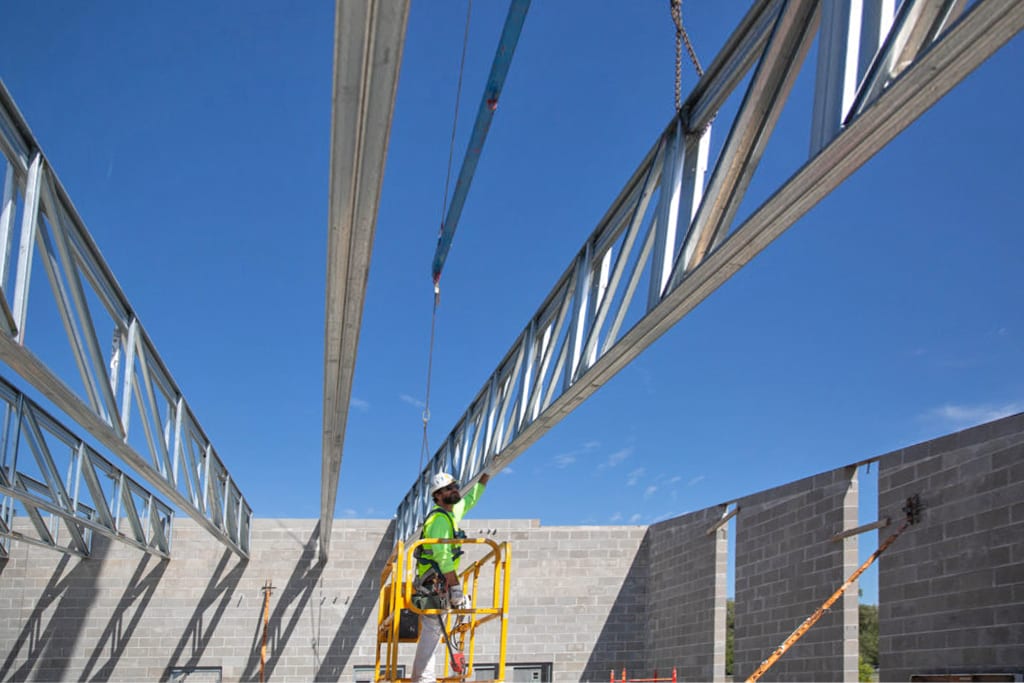 Construction worker in a lift guiding a steel truss into place on a building site under a clear sky.