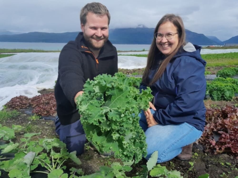 Smiling man and woman in a field holding a large kale plant, with mountains and water behind them.
