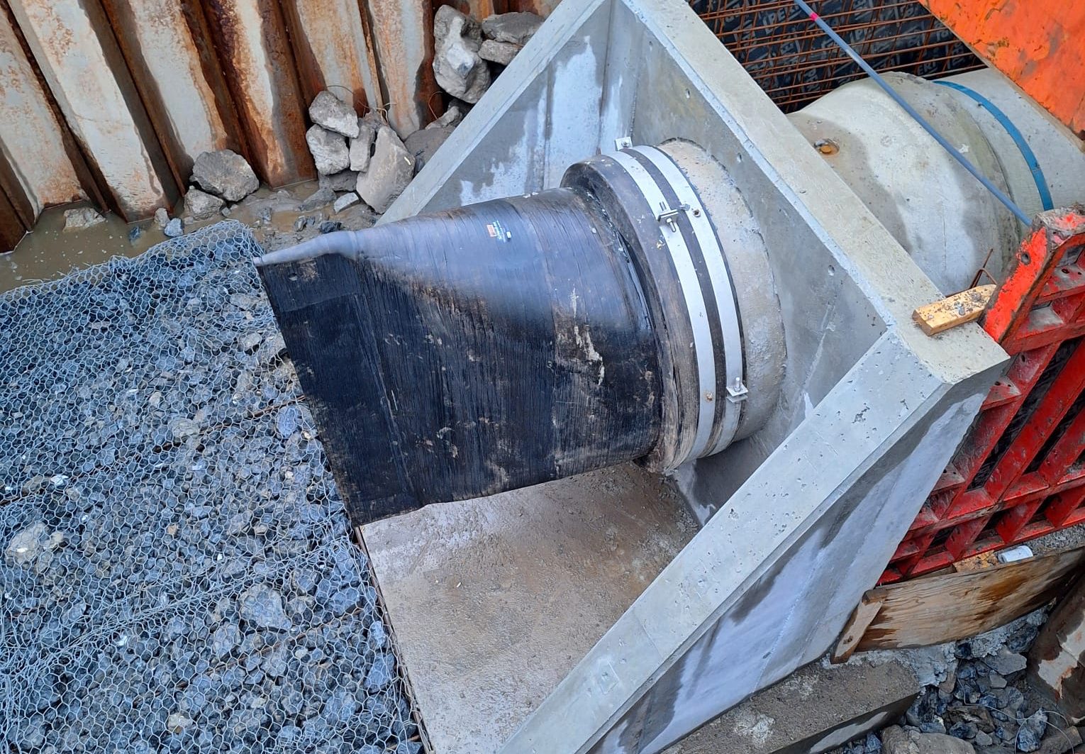 A black pipe emerges from a concrete structure, secured by metal bands, discharging onto a gabion rock mattress.