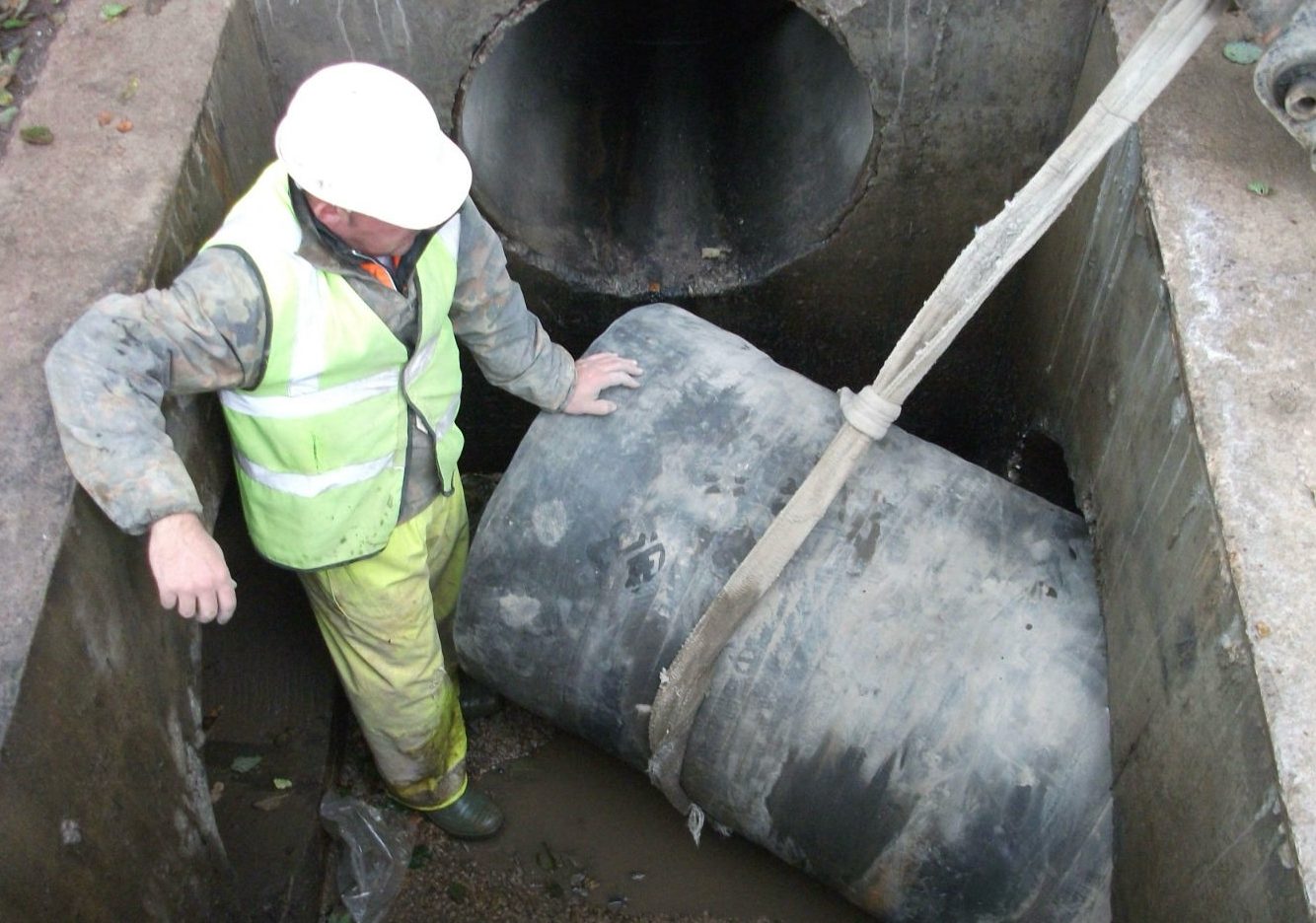 A worker in a hard hat and high-vis vest stands in a trench next to a large pipe being lowered.