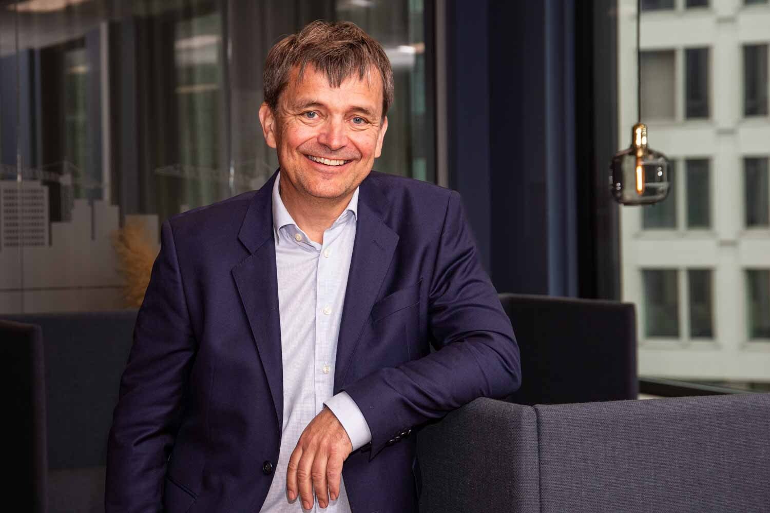 Smiling businessman in a dark blue suit and light blue shirt, leaning on a grey sofa in an office setting.
