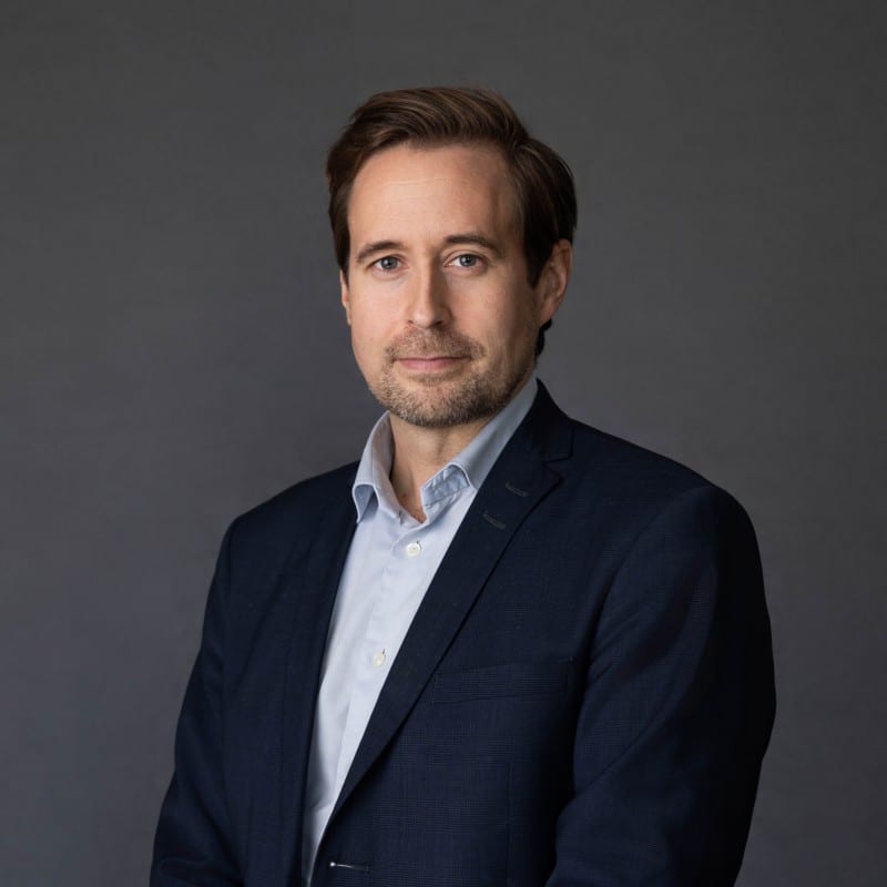 Professional headshot of a man with brown hair and light beard, wearing a dark blue suit and light shirt.